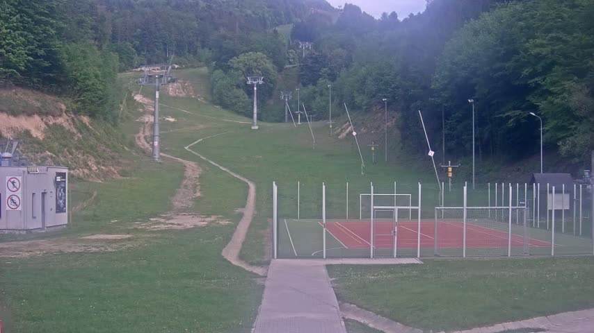 A grassy hillside with ski lifts, a paved walkway, and an enclosed multi-use sports court is visible under an overcast sky.