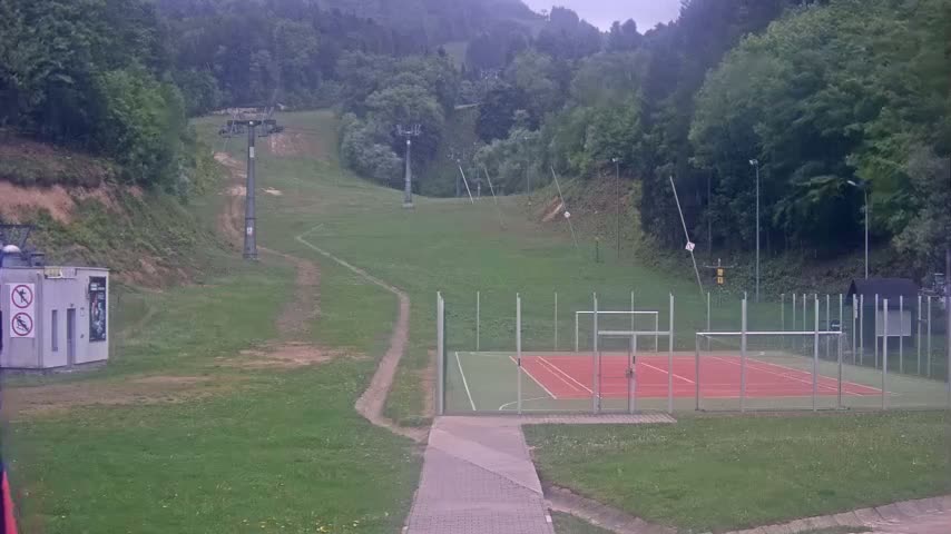 A multi-use games area with a red and green playing surface sits below a grassy hillside with a ski lift and wooded area in the background on an overcast day.