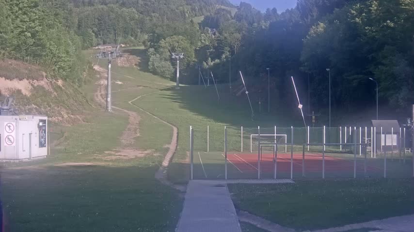 A grassy hillside with a chairlift and an enclosed sports court is visible on a sunny day.