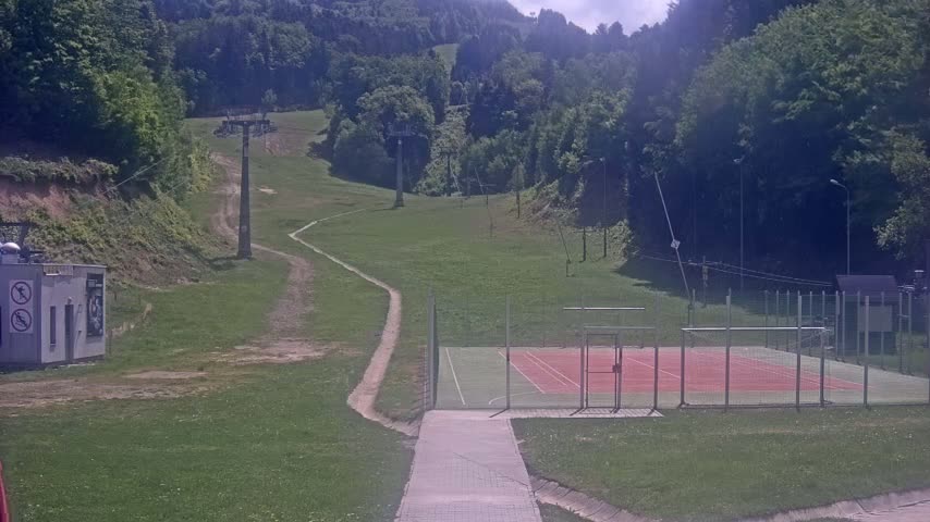 A paved pathway leads to an enclosed sports court situated on a grassy area below a ski lift and hillside covered in lush green trees under a sunny sky.