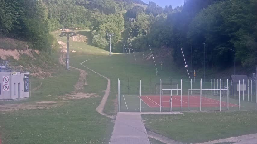 A paved pathway leads to an outdoor sports court enclosed by a fence, situated on a grassy area with a ski lift visible in the background on a partly cloudy day.