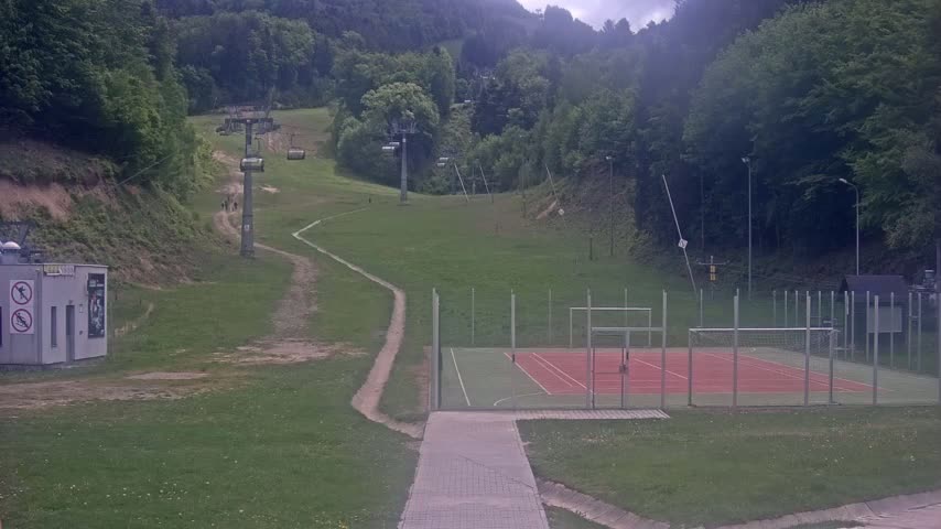A grassy hillside with a chairlift, a dirt path, and an enclosed multi-use games area is visible on an overcast day.