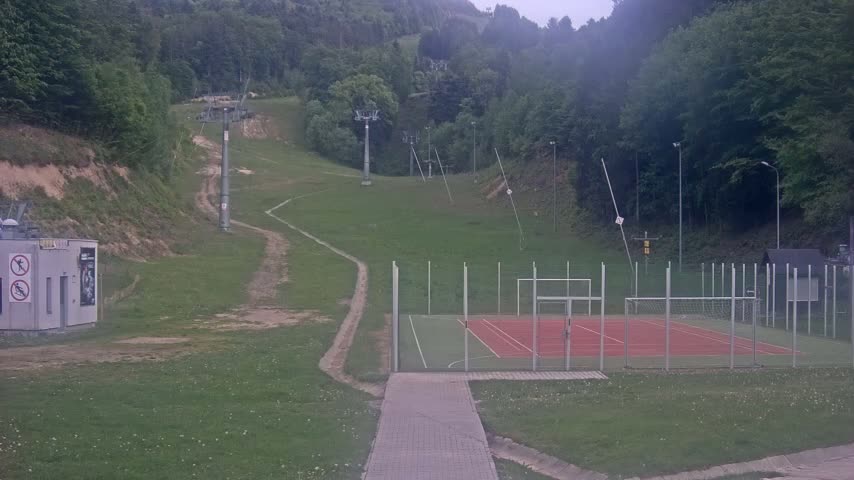 A grassy hillside with ski lift infrastructure, a small red and green sports court, and a paved walkway is visible on an overcast day.