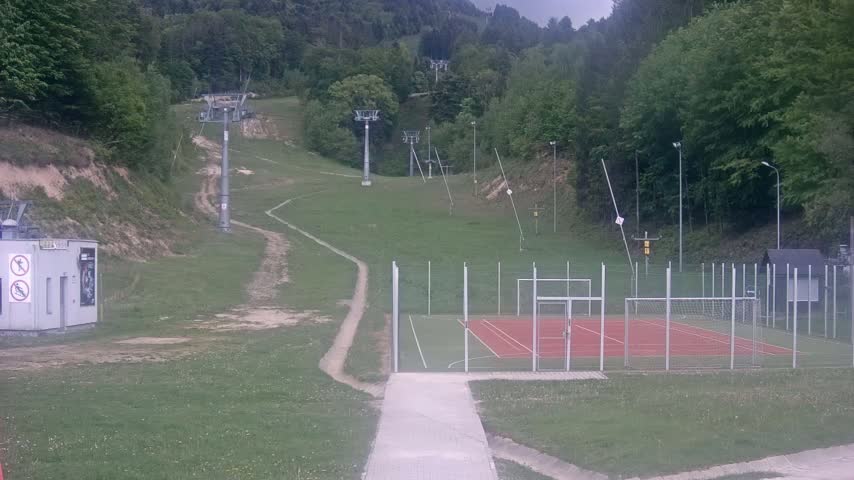 A grassy hillside with several ski lift towers, a paved walkway, and an enclosed multi-purpose sports court.