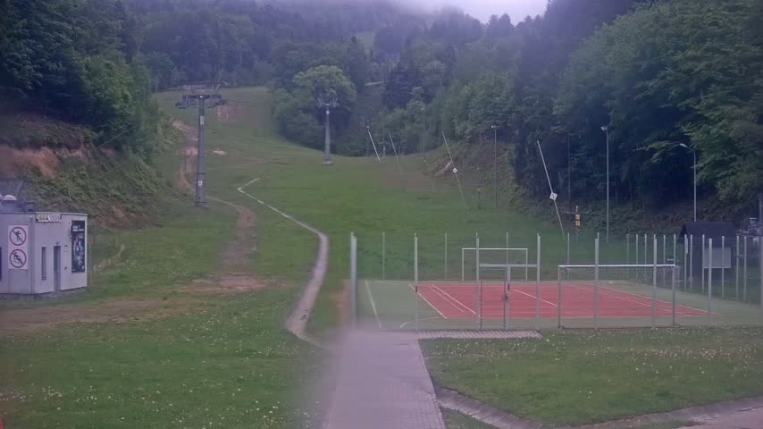 A grassy hillside with a ski lift and a small, fenced-in sports court is visible on an overcast day.