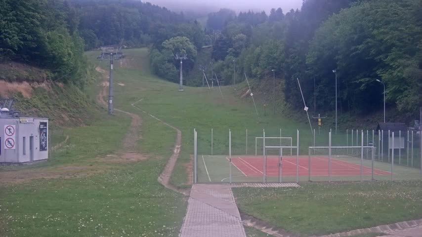 A grassy hillside with a ski lift, a small building, and an outdoor sports court is visible on an overcast day.