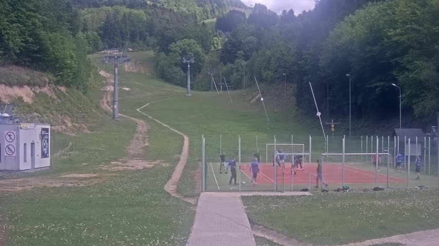 A soccer game is taking place on a small, red-colored field surrounded by grassy hills and a ski lift in an overcast setting.