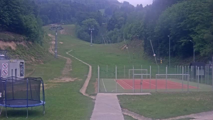 A multi-use games area with a red surface sits below a grassy hillside with a ski lift and a trampoline.