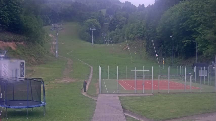 A person stands near a fenced-in sports court and trampoline at the base of a grassy hill with ski lifts and a forest in the background on an overcast day.
