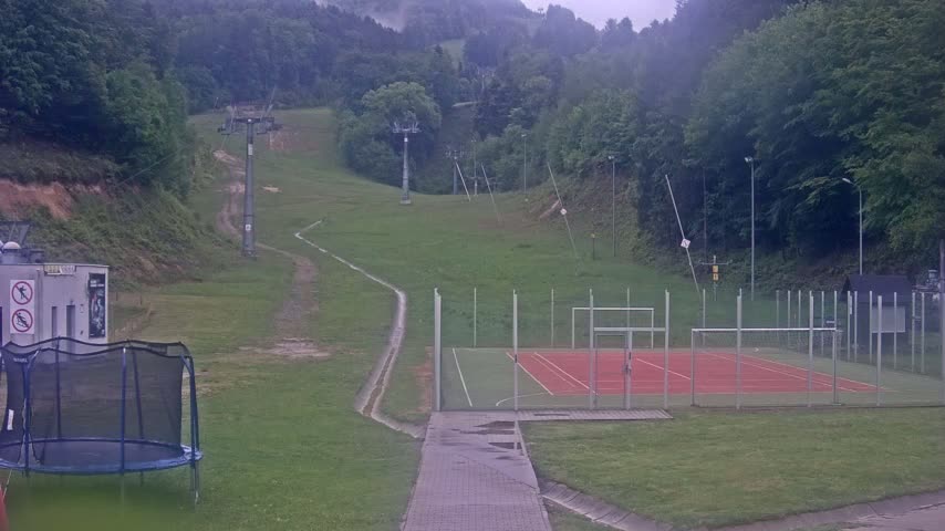 A grassy area with a ski lift in the background, a multi-purpose court, and a trampoline in the foreground, all on an overcast day.