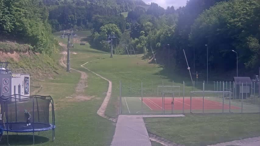 A grassy hillside with a ski lift, a playground with a trampoline, and an outdoor sports court is visible on a partly cloudy day.