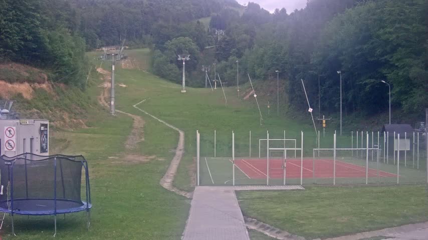 A grassy hillside with inactive ski lifts, a small multi-use sports court, and a trampoline sits below, under an overcast sky.