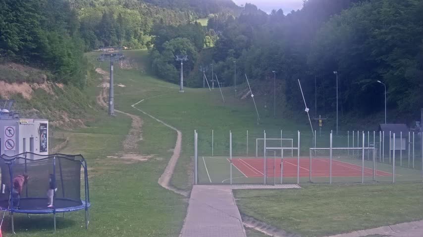 A grassy hillside with a ski lift, a small sports court, and a trampoline is visible, all under an overcast sky.