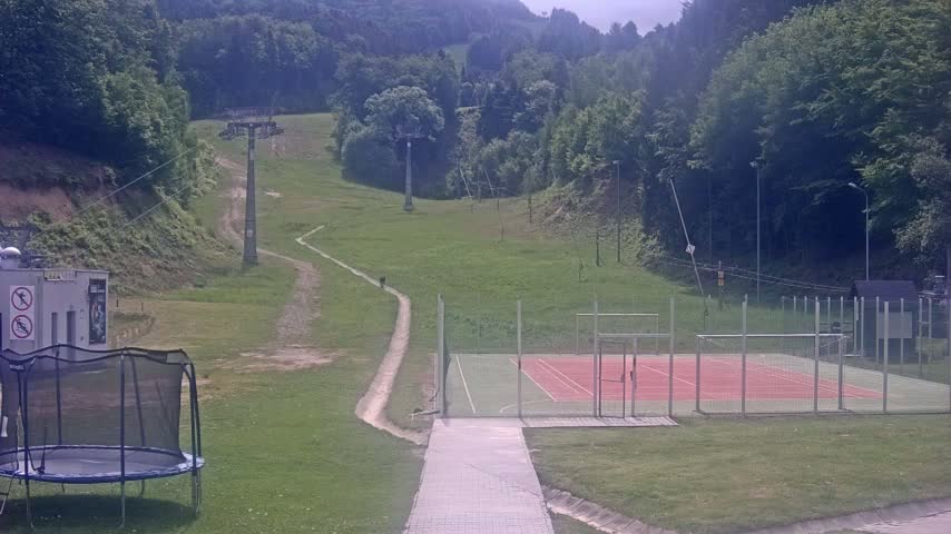 A grassy hillside with a ski lift, a paved path leading to an enclosed sports court, and a trampoline are visible on a partly cloudy day.