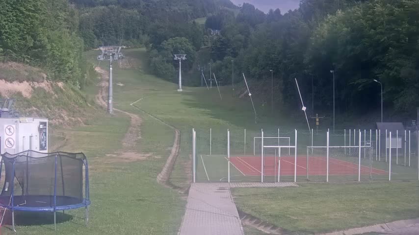 A grassy hillside with ski lift infrastructure and a multi-purpose sports court is visible, under an overcast sky.
