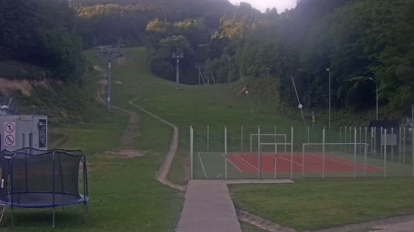 A grassy area with a small sports court, a ski lift in the background, and a trampoline is visible under an overcast sky.