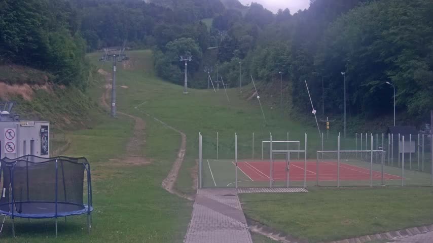 A grassy hillside with a ski lift, a small sports court, and a trampoline is visible on an overcast day.