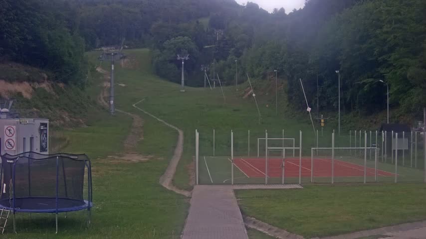 A grassy hillside with a ski lift, a small multi-use games area, and a trampoline is visible, all under an overcast sky.