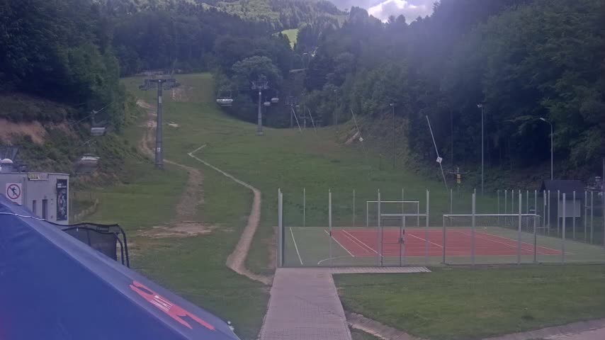 A chairlift ascends a grassy hillside dotted with trails, overlooking an enclosed multi-purpose sports court, all under a partly cloudy sky.