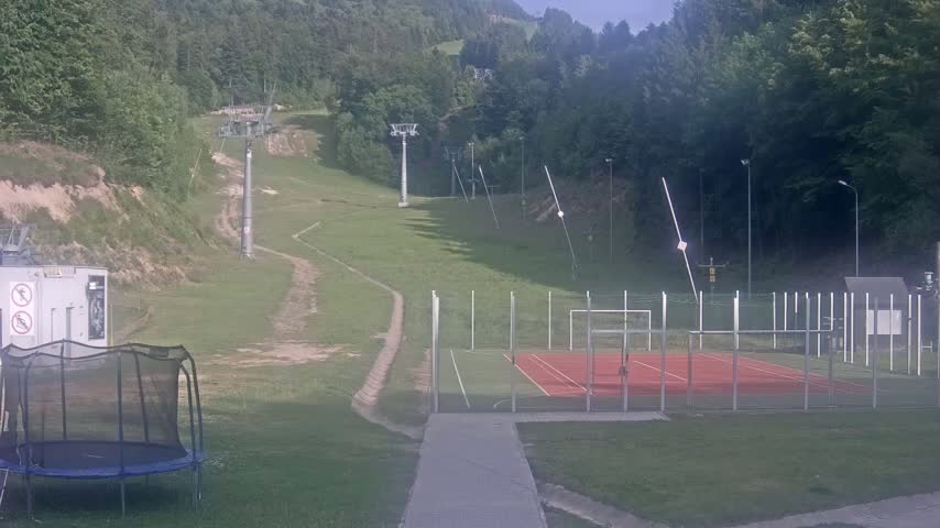 A grassy hillside with ski lifts, a fenced-in sports court, and a trampoline, all under sunny conditions.