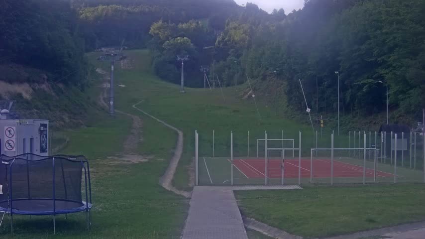 A grassy hillside with a ski lift, a small red and green sports court, and a trampoline is visible under an overcast sky.