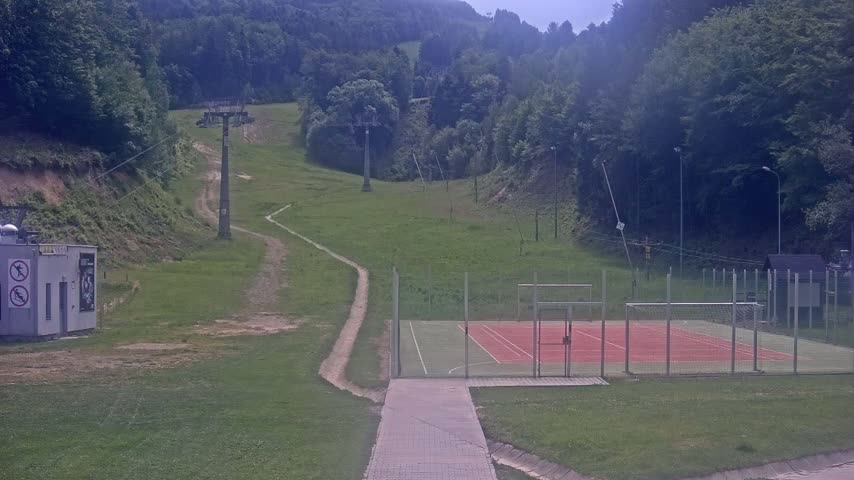 A grassy hillside with a chairlift, a paved walkway leading to an enclosed multi-use sports court, and overcast weather.