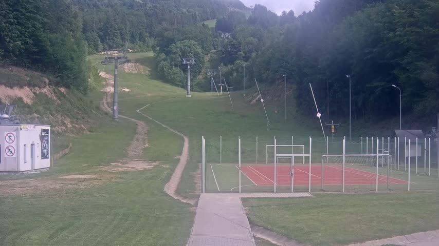 A grassy hillside with ski lifts and a small, fenced-in sports court is visible under an overcast sky.