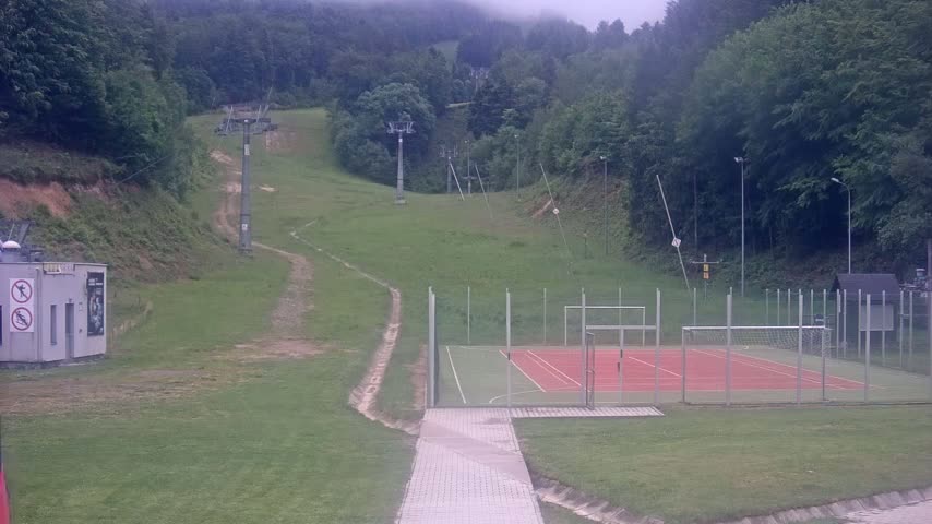 A grassy hillside with ski lift infrastructure and a fenced-in multi-purpose sports court is visible on an overcast day.