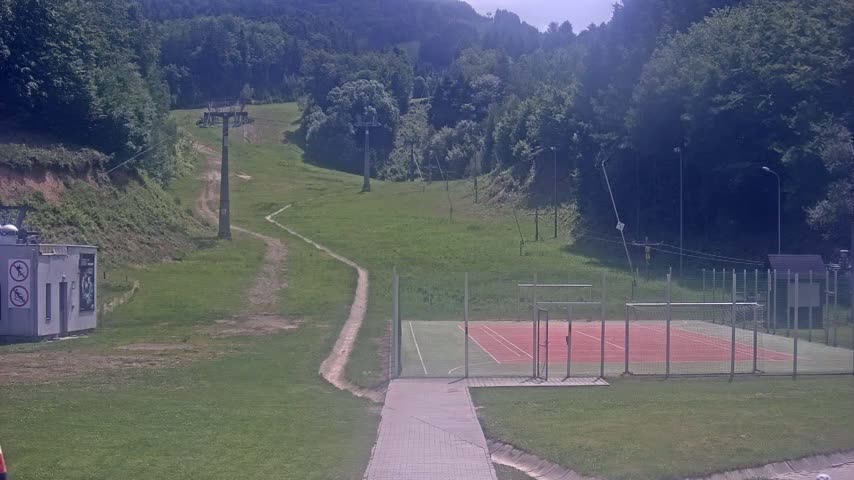 A paved walkway leads to an outdoor sports court next to a grassy hillside with a chairlift and dirt paths on a sunny day.