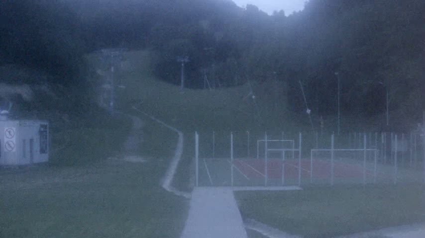 A foggy view of a paved walkway leading to an outdoor sports court, nestled among a hilly, wooded area with ski lift infrastructure visible in the background.