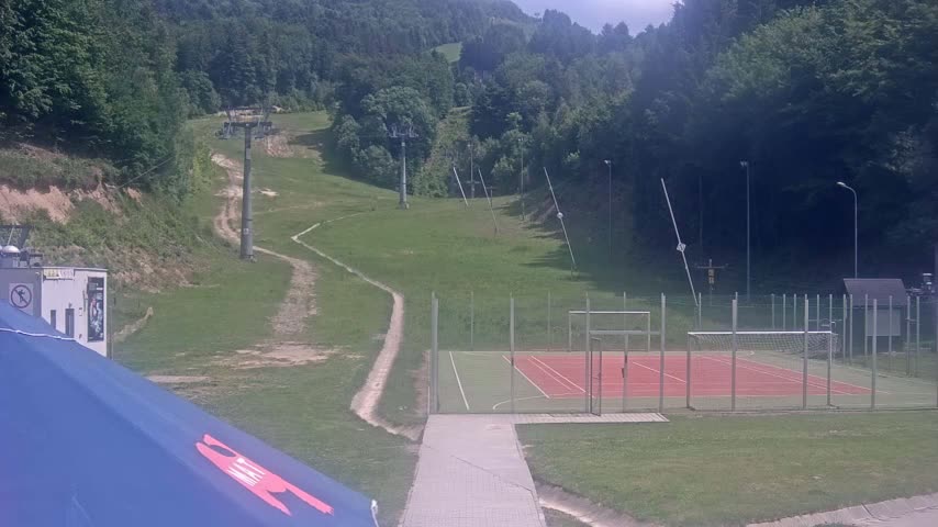 A grassy hillside with ski lifts, a dirt path, and an enclosed multi-purpose sports court is visible on a sunny day.
