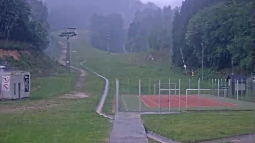 A rain-soaked hillside scene features a paved path leading to an enclosed tennis court, with a ski lift visible in the background.