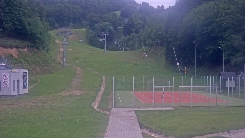 A paved walkway leads to an enclosed multi-purpose court next to a grassy hillside with a chairlift and small building in the distance under an overcast sky.