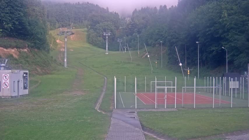 A foggy day reveals an outdoor multi-use court surrounded by a grassy area, with several ski lift towers visible on a hillside in the background.