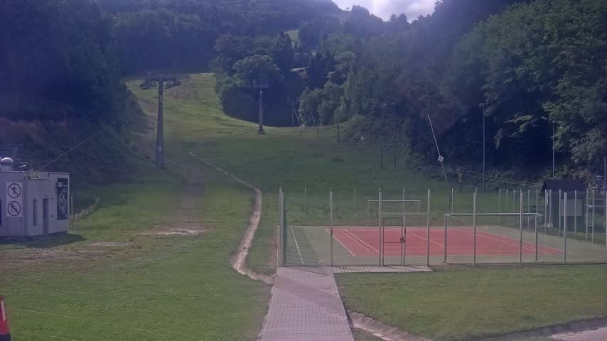 A paved walkway leads to an enclosed red sports court situated on a grassy area below a ski lift and hillside covered in trees on a partly cloudy day.