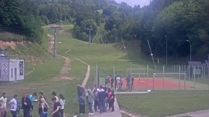 A grassy hillside with a ski lift, a small group of people gathered near a red sports court, and a building in the background, under a partly cloudy sky.