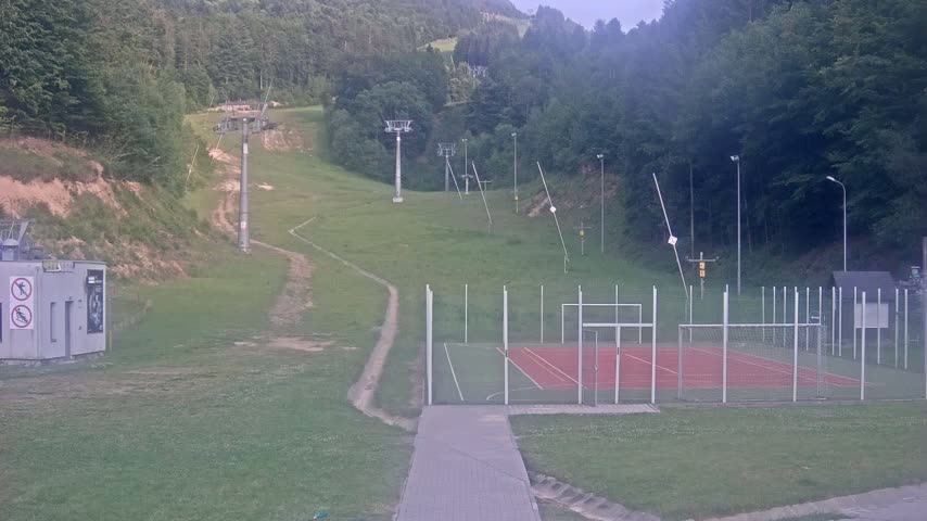 A paved walkway leads to an outdoor sports court next to a grassy hillside with ski lift infrastructure, all under an overcast sky.