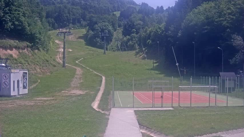 A paved pathway leads to an outdoor tennis/multi-use court surrounded by a fence, situated on a grassy hill below a ski lift and a forested mountain under sunny skies.