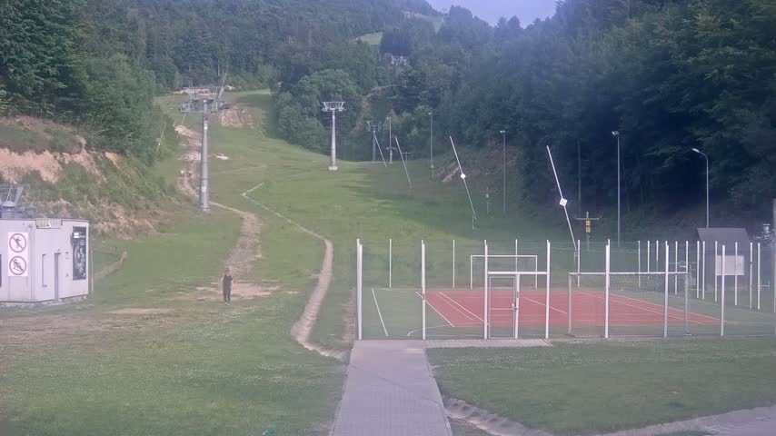 A grassy hillside with ski lift infrastructure, a small enclosed sports court, and a person walking on a path, under an overcast sky.