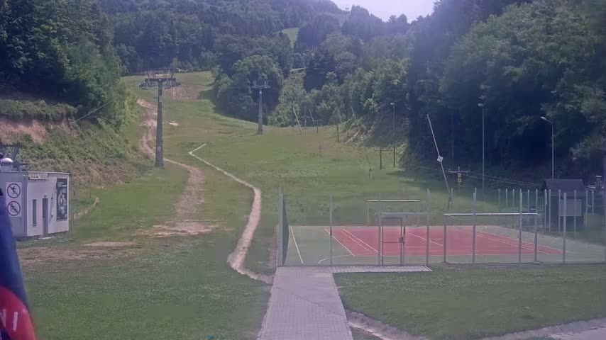 A paved pathway leads to an outdoor sports court enclosed by a fence, situated below a grassy hillside with chairlifts and lush green trees under a partly cloudy sky.