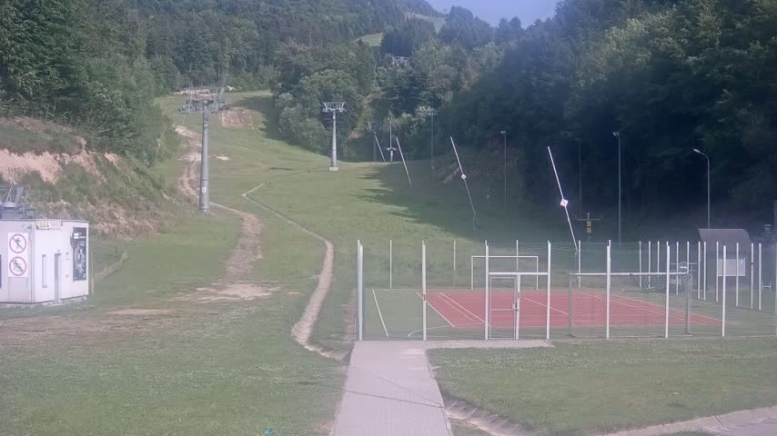 A grassy hillside with ski lifts, a paved walkway, and an enclosed multi-purpose sports court is visible on a sunny day.