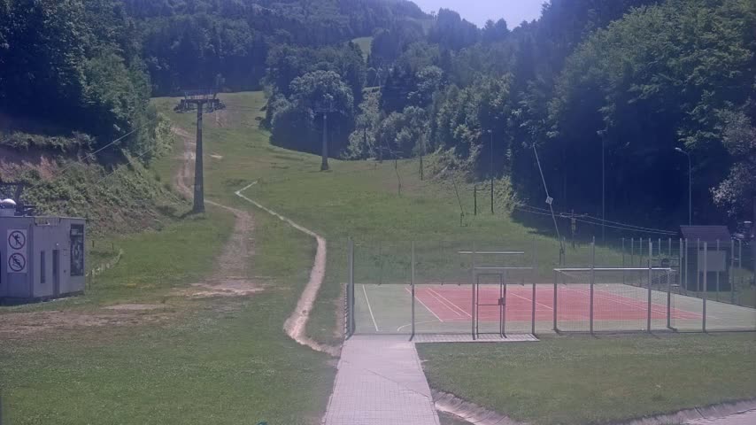 A paved walkway leads to an outdoor tennis court enclosed by a fence, situated at the base of a grassy hill with a chairlift and surrounding forest under a sunny sky.