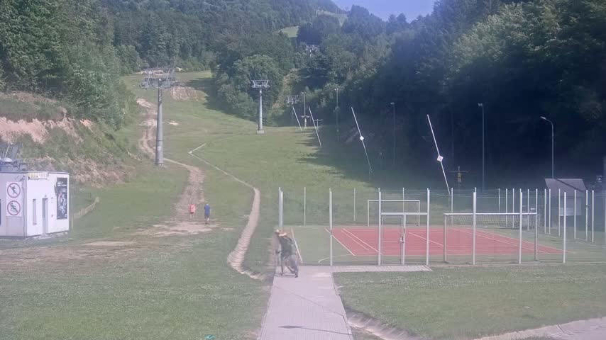 A grassy hillside with several ski lift structures, a small building, a few people walking, and a fenced-in red sports court is visible under a sunny sky.