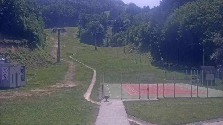 A grassy hillside with a ski lift and a tennis court is visible on a sunny day.