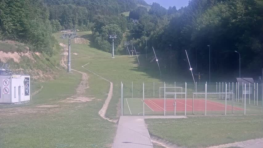 A grassy hillside with a ski lift and a fenced-in sports court is visible on a sunny day.