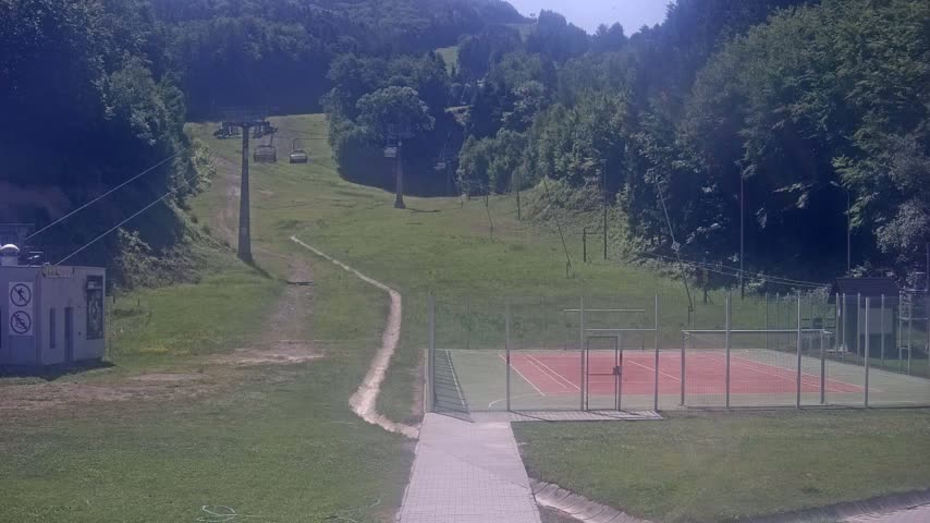 A paved walkway leads to an outdoor tennis court situated below a grassy hillside with a chairlift and surrounding forest on a sunny day.