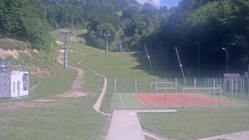 A grassy hillside with inactive ski lifts and a multi-use sports court is visible on a sunny day.