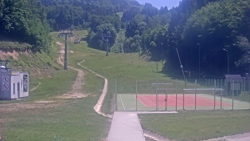 A paved walkway leads to an outdoor tennis/futsal court situated below a grassy hill with ski lifts and a wooded mountain in the background on a sunny day.