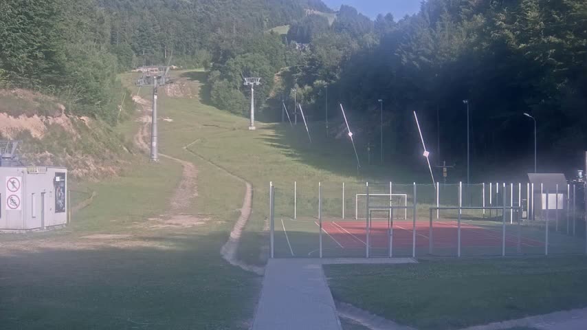 A grassy hillside with ski lift infrastructure and a fenced-in sports court is visible on a sunny day.