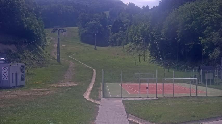 A paved walkway leads to an enclosed sports court next to a grassy hillside with a chairlift and trails.